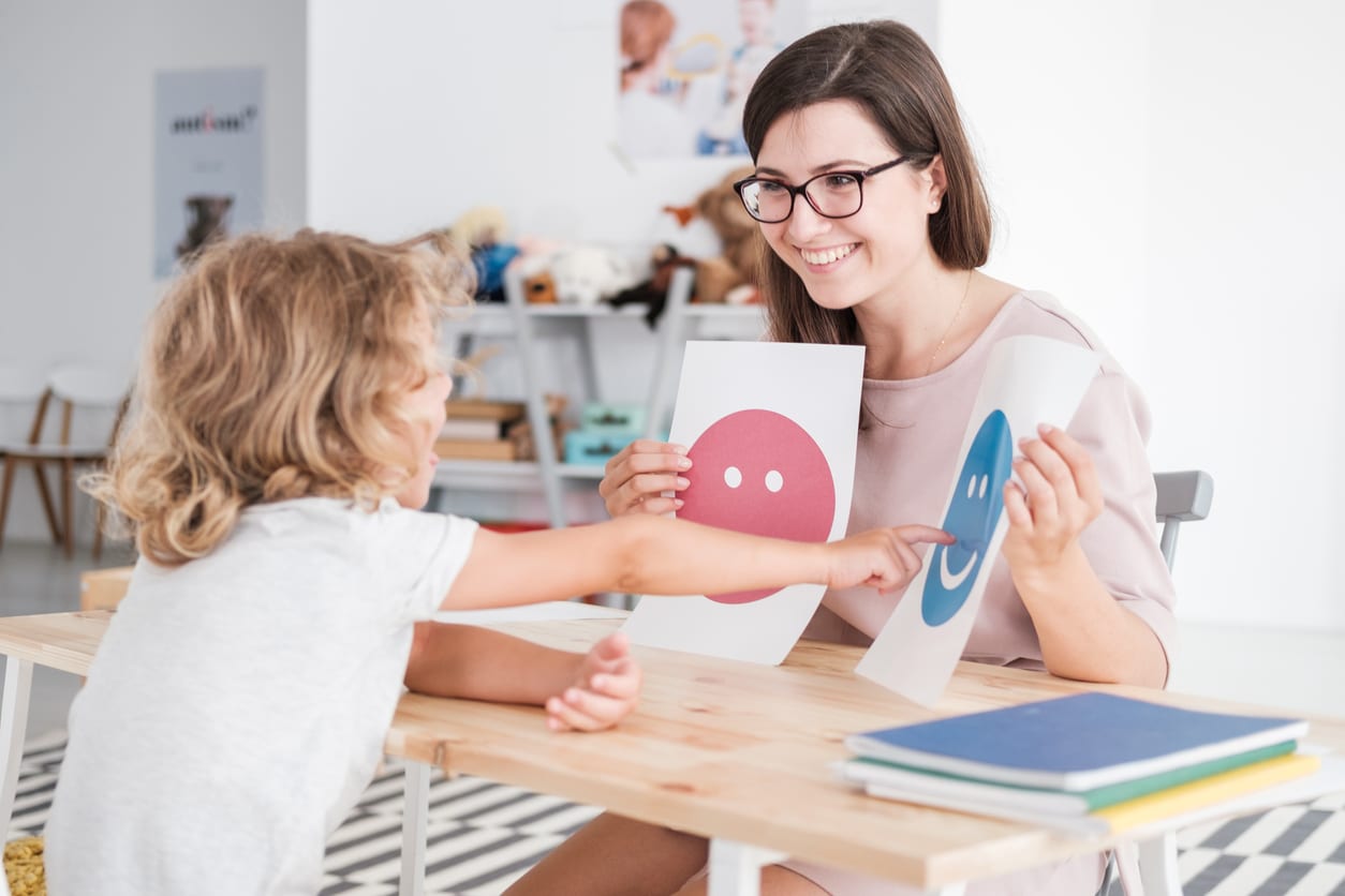 Child sitting at a table with an female RBT pointing at a picture of a sad face which is being held up by the RBT